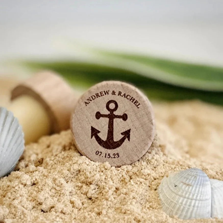 A personalized wooden wine bottle stopper engraved with a couple's names and wedding date nestled in beach sand beside white seashells