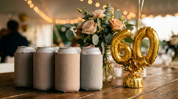 An elegant 60th birthday party table with personalized can-cooler koozies, gold number-60 balloons, and blush pink roses at golden hour